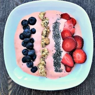 a white bowl filled with strawberry protein smoothie bowl and topped with blueberries, granola, chia seeds, and sliced strawberries