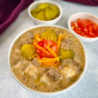 slow cooker cheeseburger soup in a white bowl with shredded cheese, pickles, and tomatoes