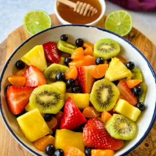 fresh strawberries, mandarin oranges, kiwi, blueberries, and pineapple fruit salad in a white bowl with honey lime dressing