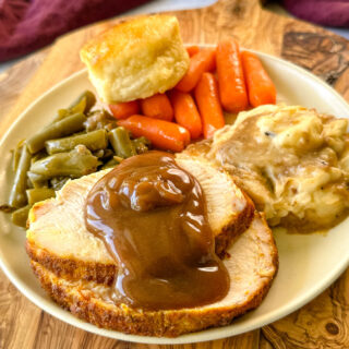 boneless turkey breast slices on a plate with mashed potatoes, gravy, green beans, carrots, and a biscuit