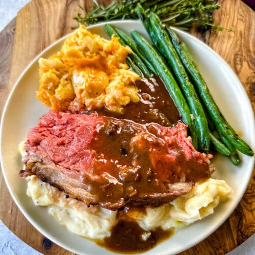 garlic herb butter roasted prime rib with au jus gravy on a plate with green beans, mashed potatoes, and mac and cheese