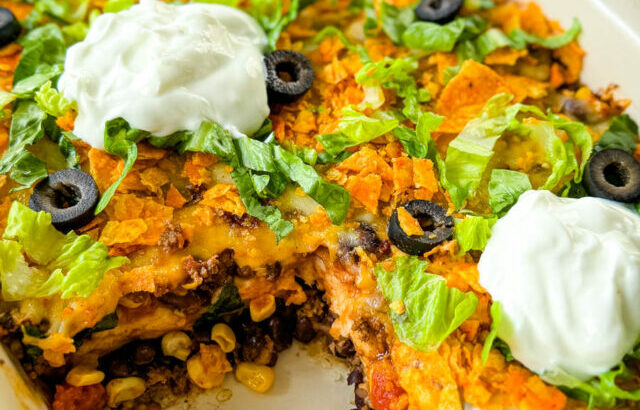 taco bake caserole in a baking dish with Doritos, olives, lettuce, and sour cream