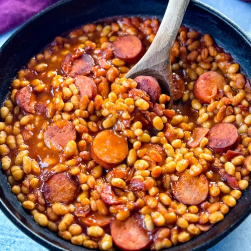 homemade pork and beans with sausage, bacon, and a wooden spoon in a cast iron skillet