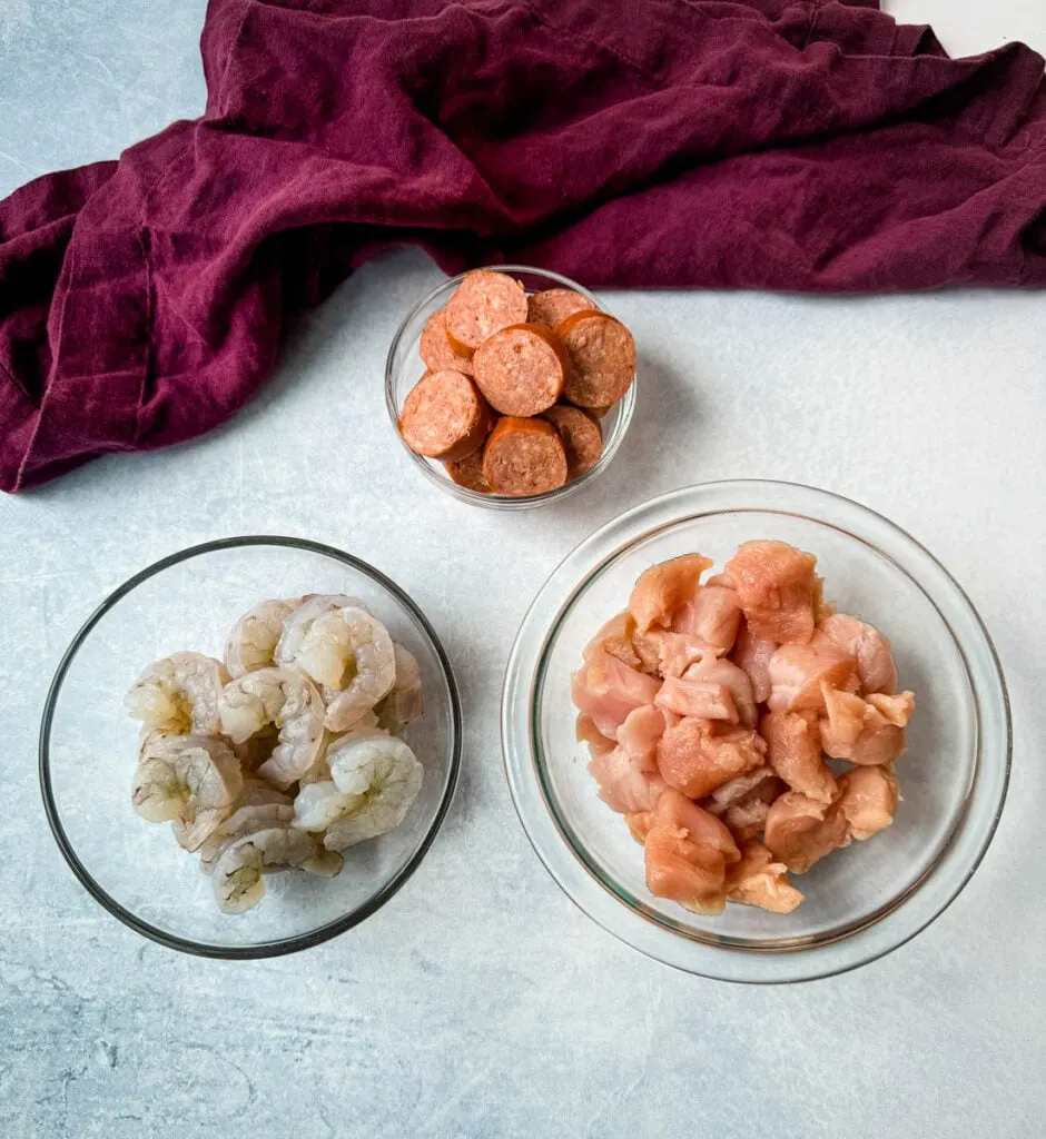 smoked sausage, raw shrimp, and raw chicken thighs in separate glass bowls