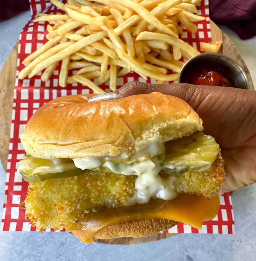 person holding fish sandwich with cheese, pickles, and tartar sauce on a brioche bun on a plate with fries