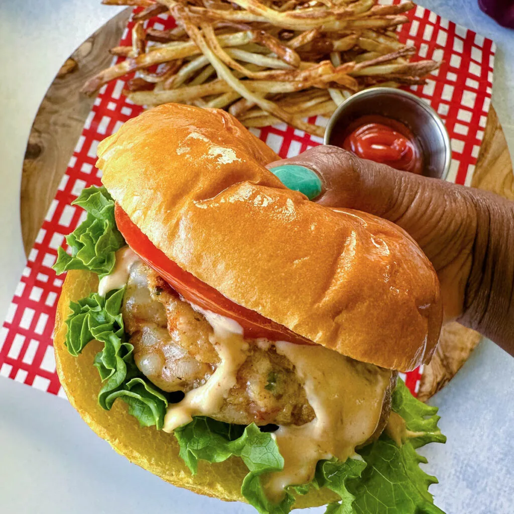 person holding shrimp burger with lettuce, tomato, and spicy mayo