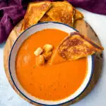 homemade creamy tomato soup in a white bowl with croutons with slices of grilled cheese on a flat surface