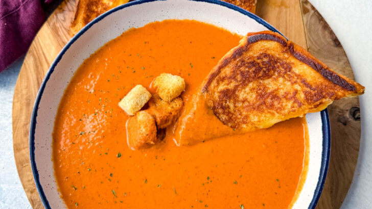 homemade creamy tomato soup in a white bowl with croutons with slices of grilled cheese on a flat surface