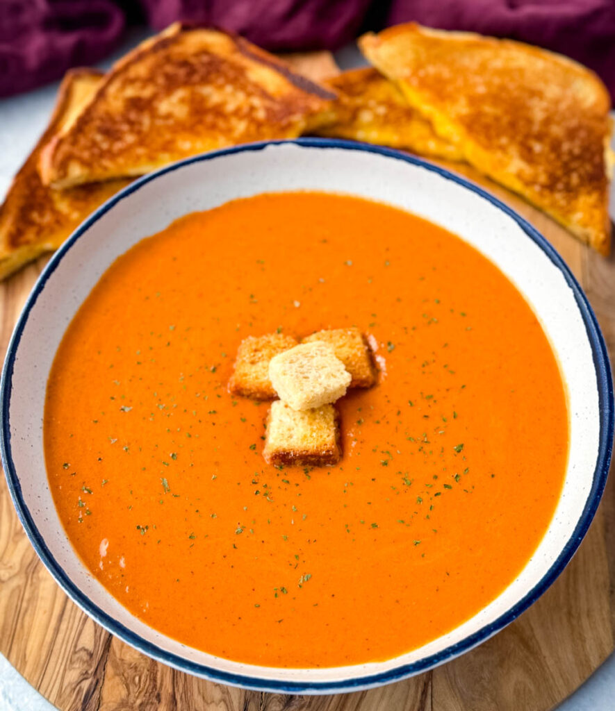 homemade creamy tomato soup in a white bowl with croutons with slices of grilled cheese on a flat surface