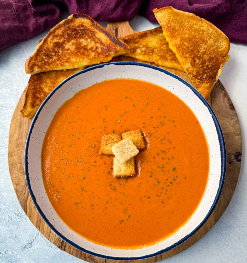 homemade creamy tomato soup in a white bowl with croutons with slices of grilled cheese on a flat surface