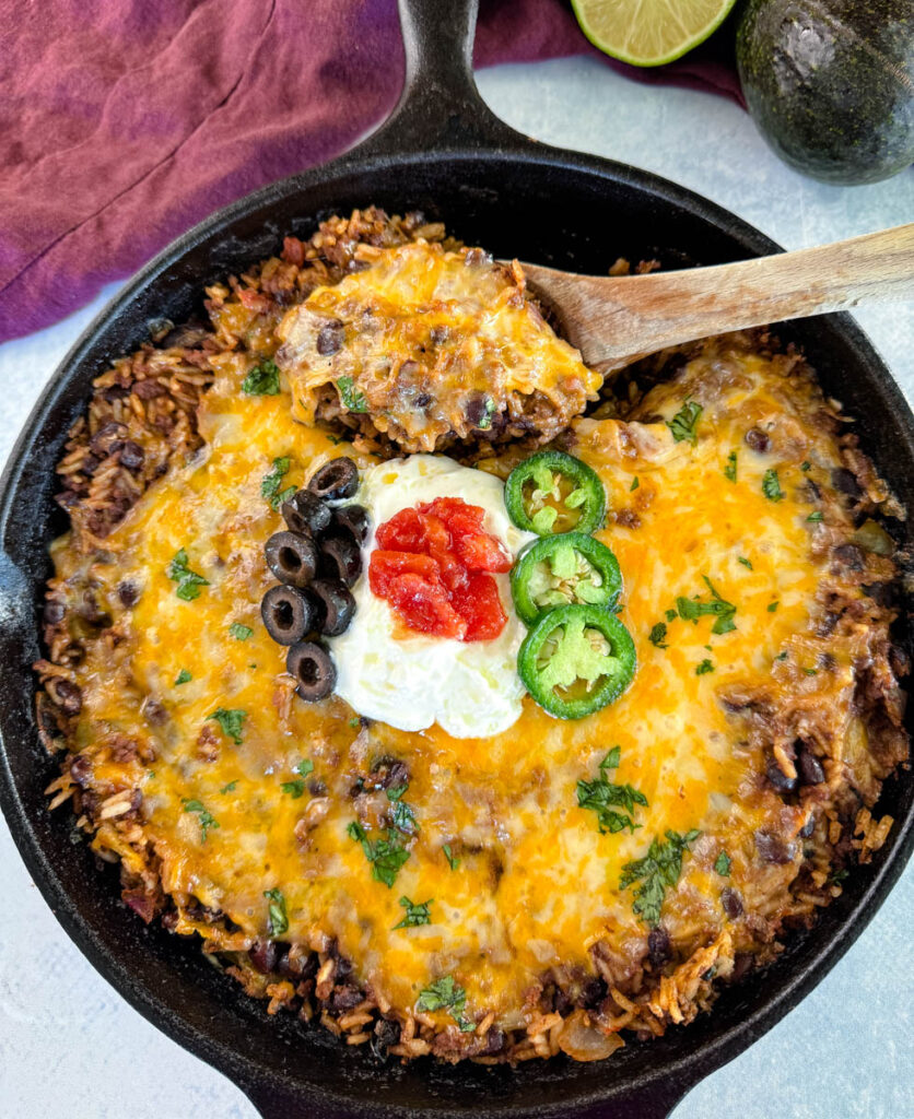 Mexican beef and rice taco skillet dish in a cast iron skillet with a wooden spoon and sour cream, tomatoes, and black olives