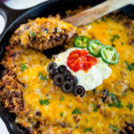 Mexican beef and rice taco skillet dish in a cast iron skillet with a wooden spoon and sour cream, tomatoes, and black olives