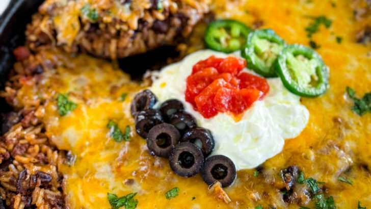 Mexican beef and rice taco skillet dish in a cast iron skillet with a wooden spoon and sour cream, tomatoes, and black olives