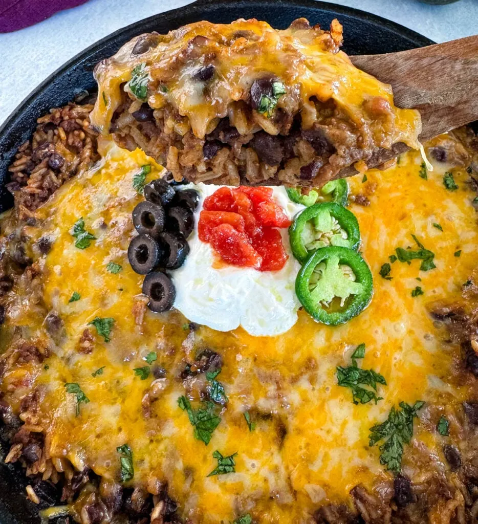 Mexican beef and rice taco skillet dish in a cast iron skillet with a wooden spoon and sour cream, tomatoes, and black olives