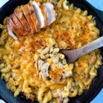 chicken mac and cheese in a cast iron skillet with a wooden spoon and sliced chicken