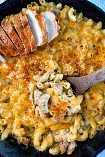 chicken mac and cheese in a cast iron skillet with a wooden spoon and sliced chicken