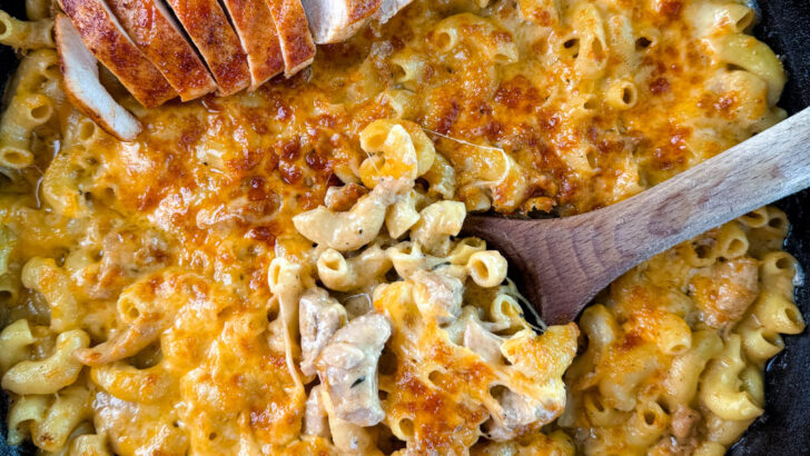 chicken mac and cheese in a cast iron skillet with a wooden spoon and sliced chicken