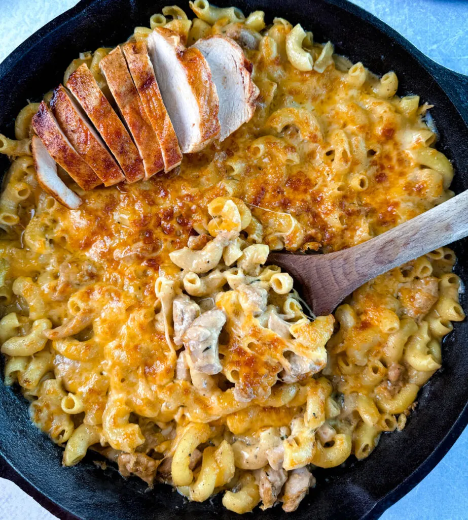 chicken mac and cheese in a cast iron skillet with a wooden spoon and sliced chicken