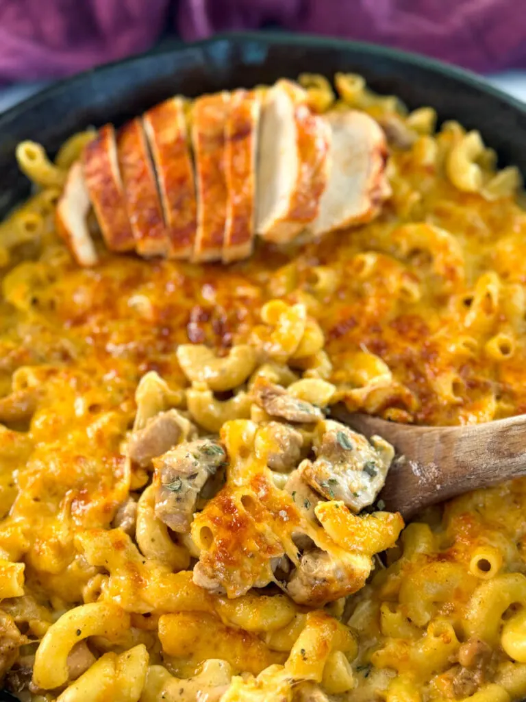 chicken mac and cheese in a cast iron skillet with a wooden spoon and sliced chicken