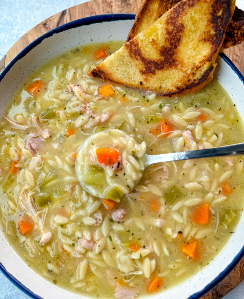 chicken orzo soup with carrots, celery, and a wooden spoon in a white bowl with a piece of bread and a spoon