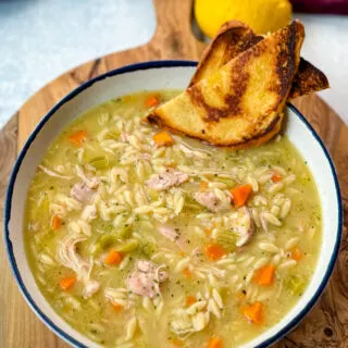 chicken orzo soup with carrots, celery, and a wooden spoon in a white bowl with a piece of bread