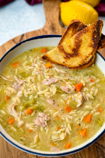 chicken orzo soup with carrots, celery, and a wooden spoon in a white bowl with a piece of bread