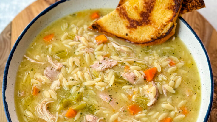 chicken orzo soup with carrots, celery, and a wooden spoon in a white bowl with a piece of bread