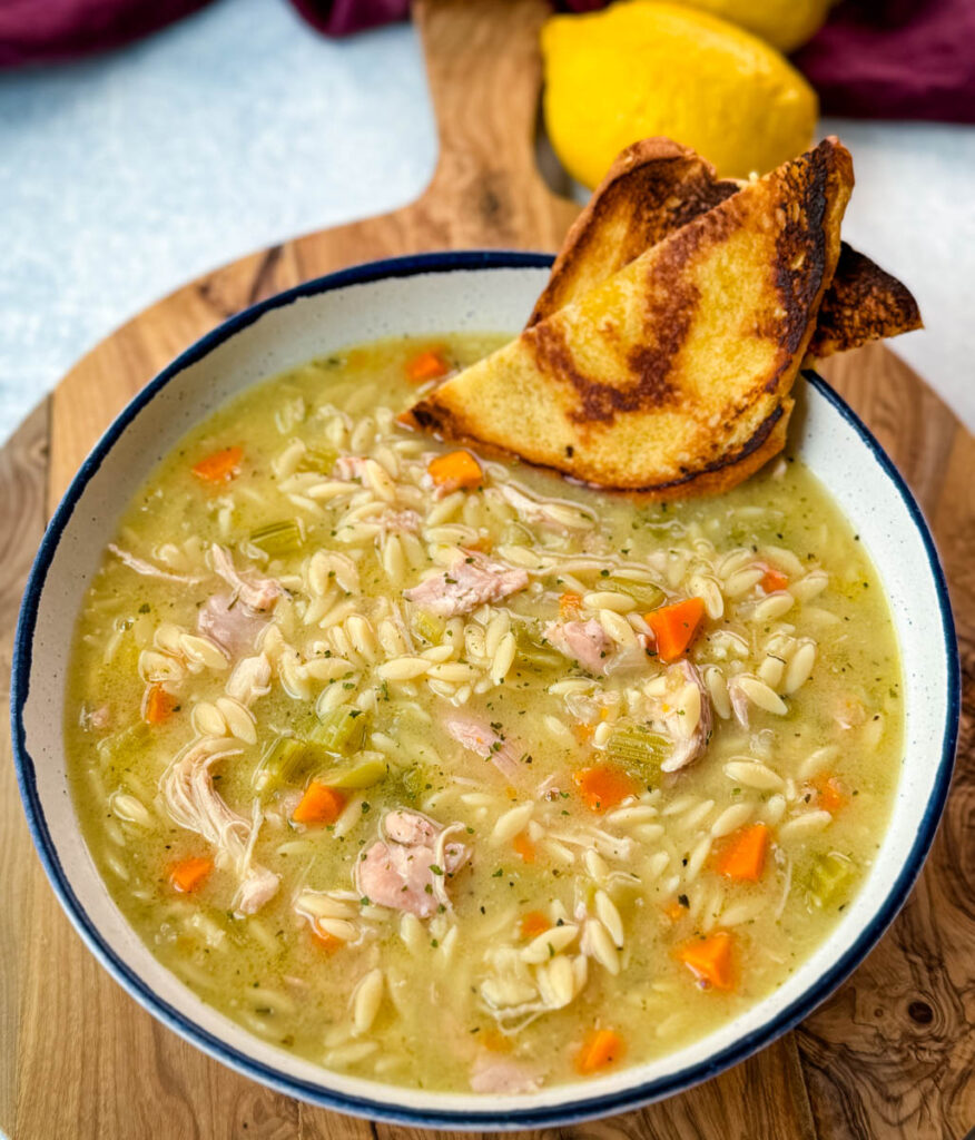 chicken orzo soup with carrots, celery, and a wooden spoon in a white bowl with a piece of bread