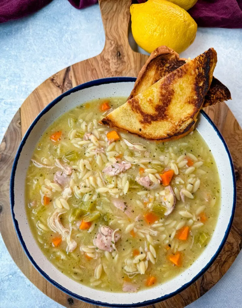 chicken orzo soup with carrots, celery, and a wooden spoon in a white bowl with a piece of bread