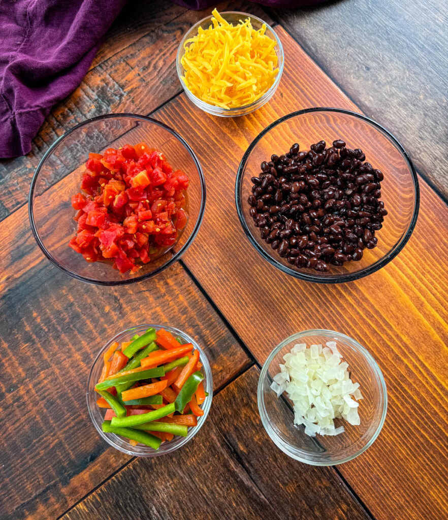 grated cheddar cheese, diced tomatoes, black beans, sliced bell peppers, and diced onions in separate bowls