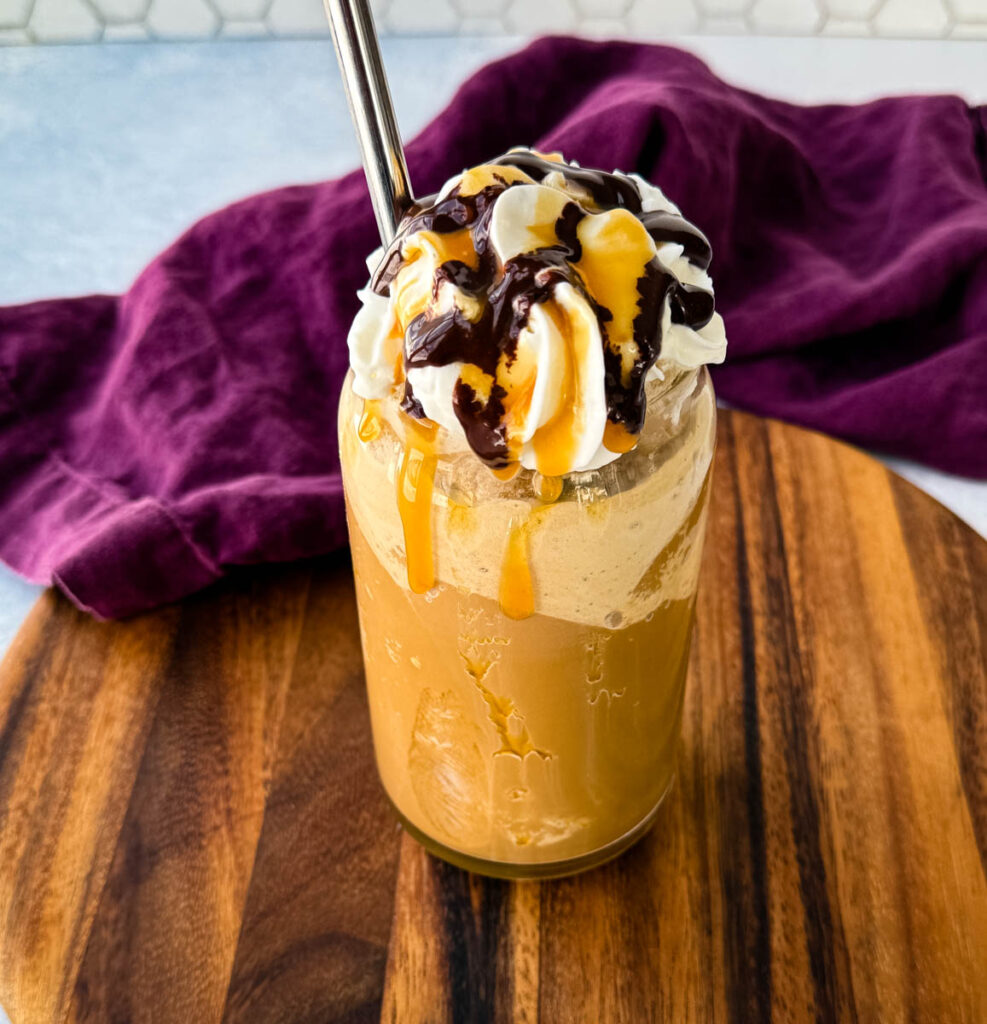 A tall glass filled with a blended coffee frappe, topped with whipped cream, chocolate drizzle, and caramel sauce. A metal straw is inserted, and the glass sits on a wooden board with a purple cloth in the background.