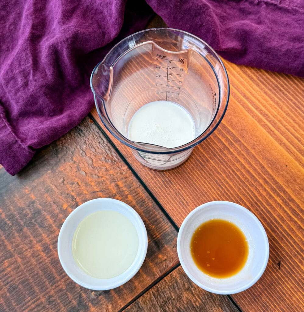 A small measuring cup containing milk and two small bowls with different liquid ingredients—one vanilla extract and sweetener—placed on a wooden table with a purple cloth in the background.