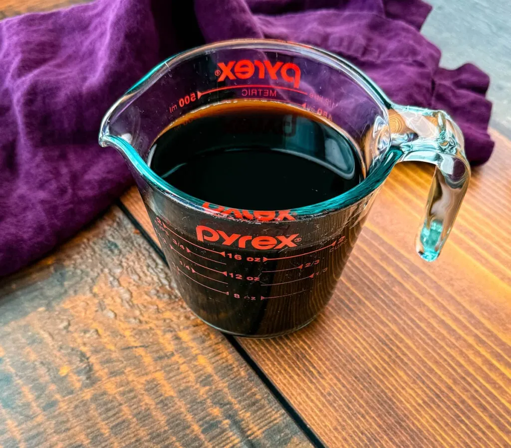 A glass Pyrex measuring cup filled with freshly brewed black coffee, sitting on a wooden table with a purple cloth partially draped behind it.