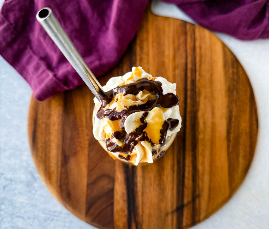 A top-down view of a coffee frappe in a glass, covered with whipped cream, chocolate drizzle, and caramel sauce. A metal straw is inserted, and the drink is placed on a wooden board with a purple cloth partially visible.