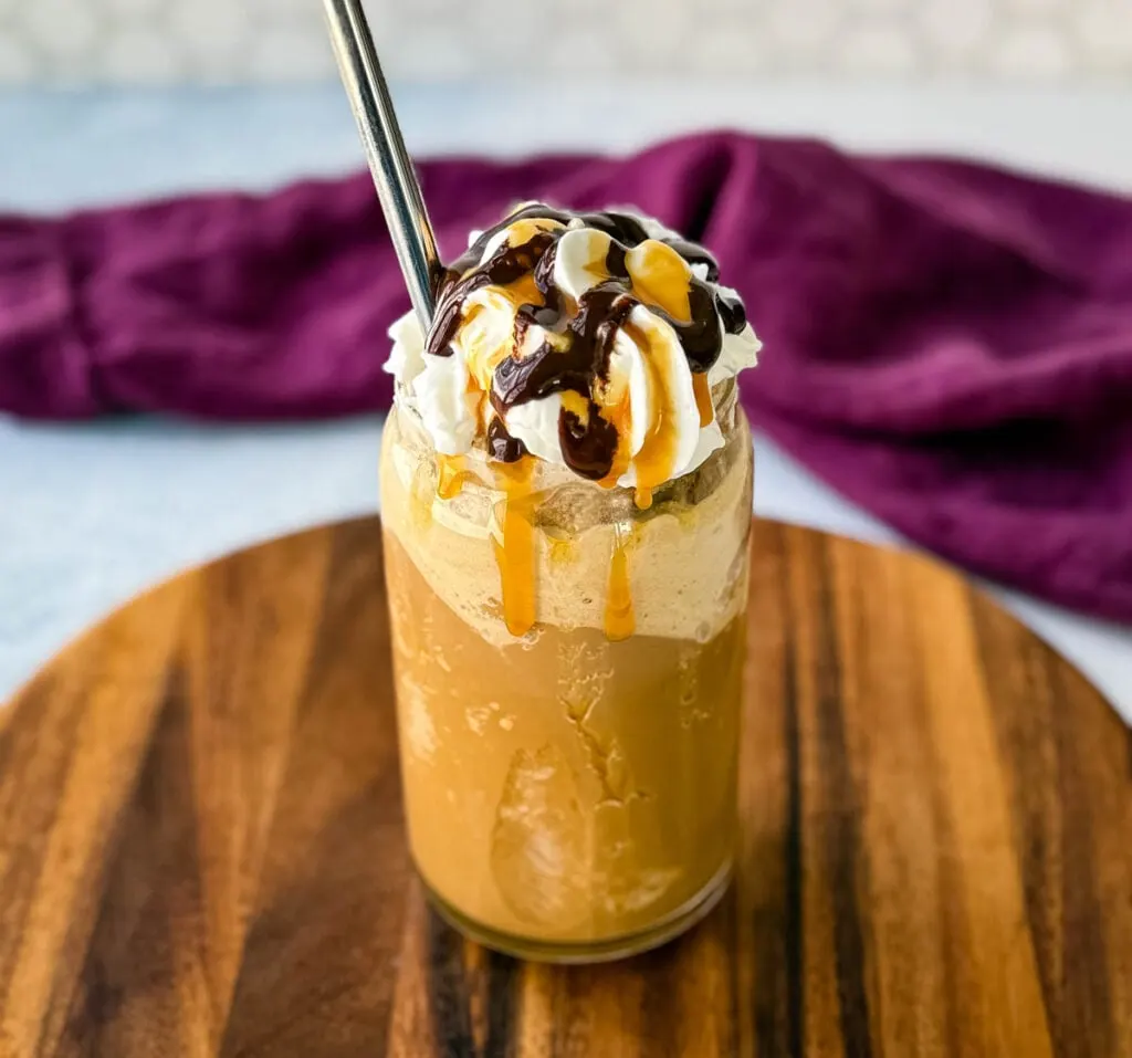 A tall glass filled with a blended coffee frappe, topped with whipped cream, chocolate drizzle, and caramel sauce. A metal straw is inserted, and the glass sits on a wooden board with a purple cloth in the background.