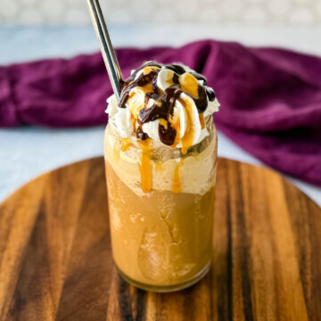 A tall glass filled with a blended coffee frappe, topped with whipped cream, chocolate drizzle, and caramel sauce. A metal straw is inserted, and the glass sits on a wooden board with a purple cloth in the background.