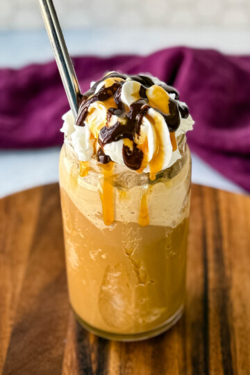 A tall glass filled with a blended coffee frappe, topped with whipped cream, chocolate drizzle, and caramel sauce. A metal straw is inserted, and the glass sits on a wooden board with a purple cloth in the background.