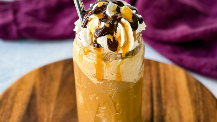 A tall glass filled with a blended coffee frappe, topped with whipped cream, chocolate drizzle, and caramel sauce. A metal straw is inserted, and the glass sits on a wooden board with a purple cloth in the background.