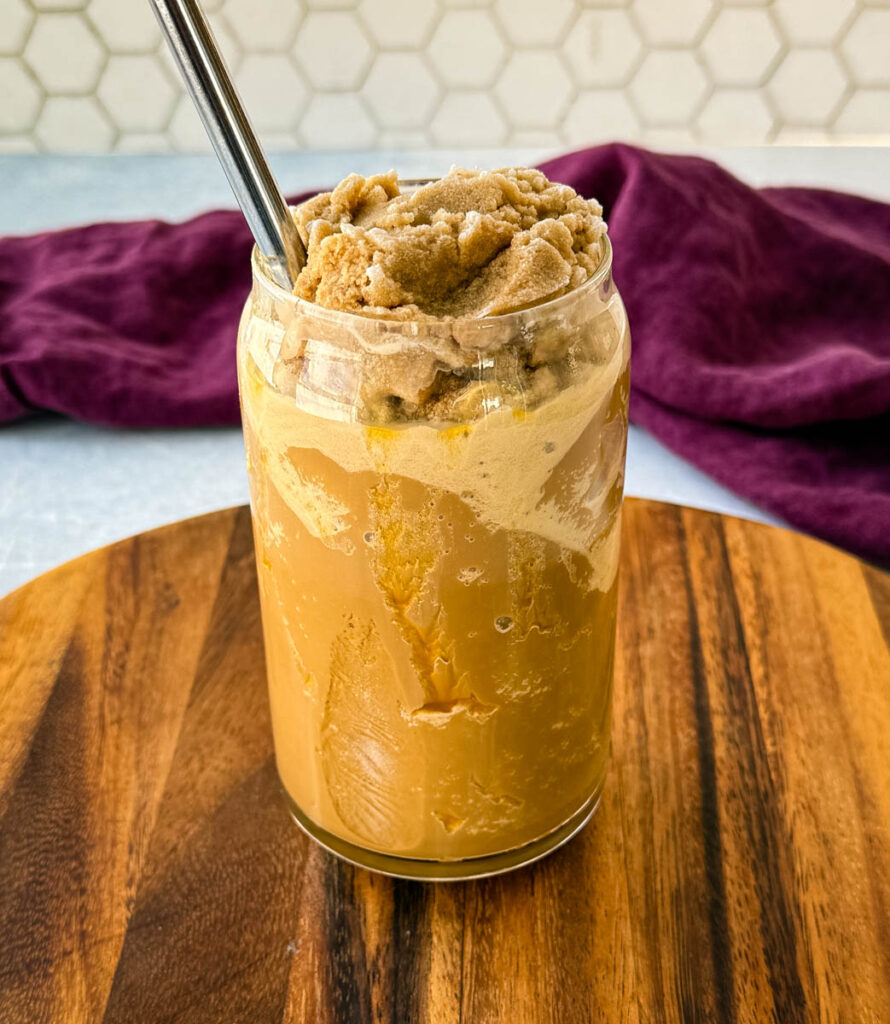 A glass filled with a blended coffee frappe topped with a thick, icy coffee mixture. A metal straw is inserted, and the glass sits on a wooden board with a purple cloth in the background.