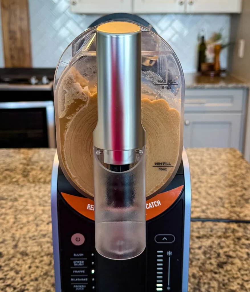 A close-up of a Ninja slushie machine filled with thick, blended coffee frappe. The dispensing spout is visible, and the machine is set against a kitchen backdrop with white cabinets and a tiled backsplash.