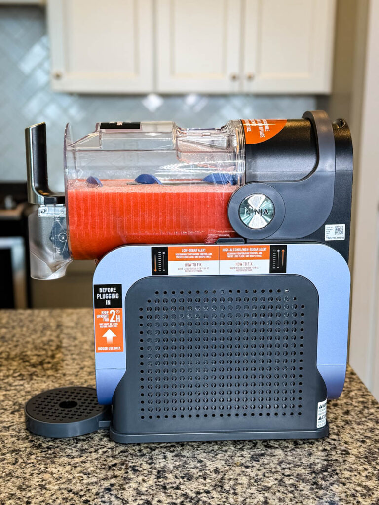 Full Side View of the Ninja SLUSHi Machine on a Kitchen Counter – The machine is placed on a kitchen counter with the chamber filled with a frozen drink. Labels on the side provide usage instructions, including a reminder to keep the unit upright for 2 hours before first use.