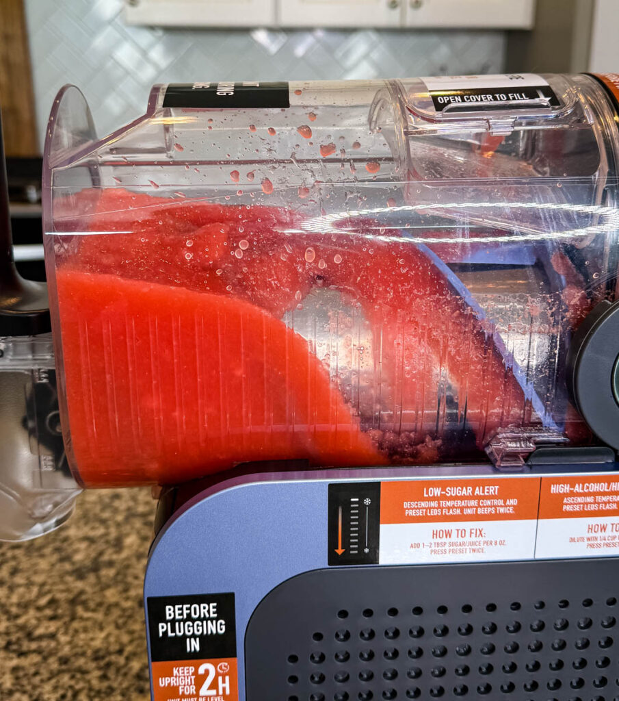 Side View of the Ninja SLUSHi Machine in Action – A side angle of the machine with red slush forming inside the chamber. The condensation and mixing process are visible, along with labels for low-sugar and high-alcohol warnings.