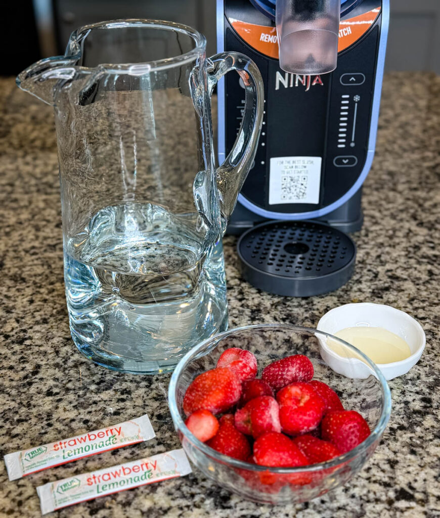 A glass pitcher of water, frozen strawberries, strawberry lemonade drink mix packets, and simple syrup laid out on a granite countertop.