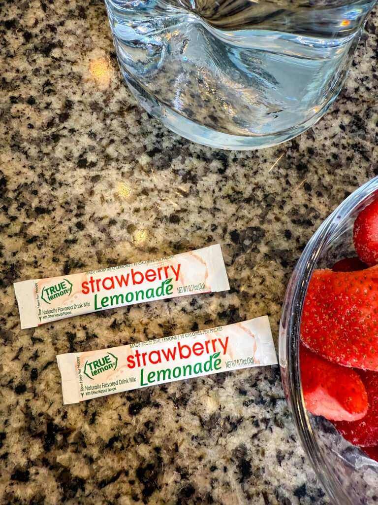 Two True Lemon Strawberry Lemonade drink mix packets on a countertop next to fresh ingredients.