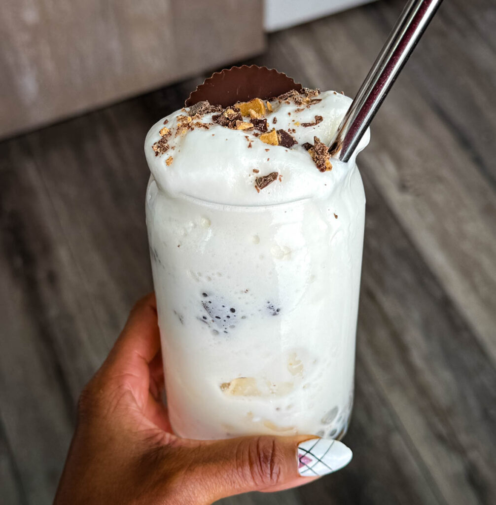 person holding Finished Ninja SLUSHi Peanut Butter Cup Vanilla Protein Shake in a glass, topped with whipped cream, crushed peanut butter cup pieces, and a whole peanut butter cup, with a metal straw for serving.