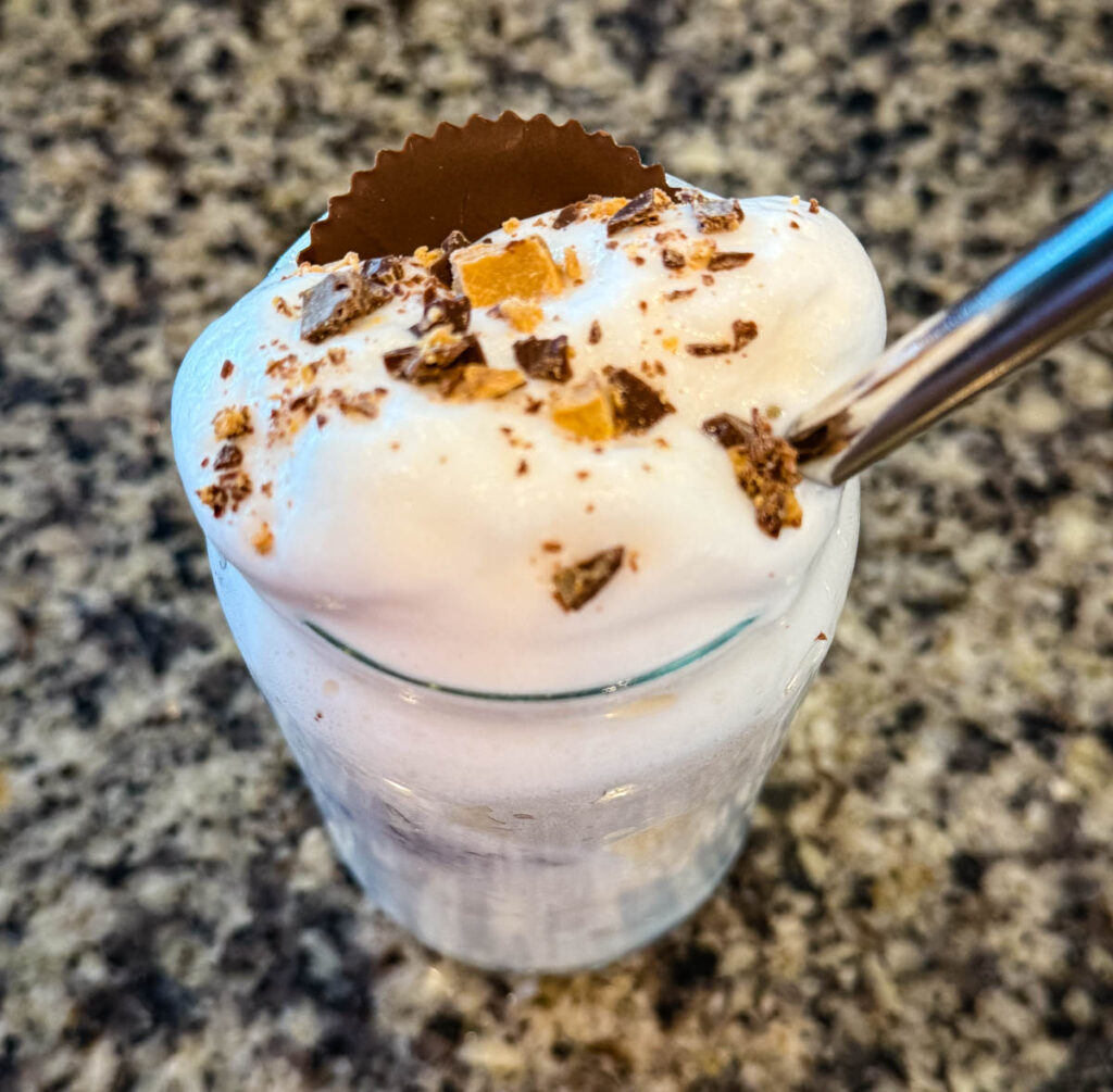 Finished Ninja SLUSHi Peanut Butter Cup Vanilla Protein Shake in a glass, topped with whipped cream, crushed peanut butter cup pieces, and a whole peanut butter cup, with a metal straw for serving.