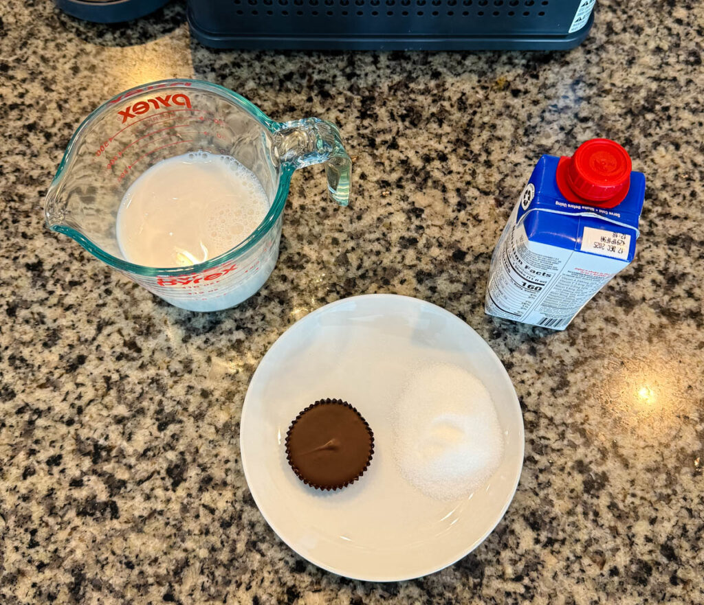 Ingredients for the Ninja SLUSHi Vanilla Protein Milkshake, including milk, sugar, a liquid vanilla protein shake, and a protein peanut butter cup displayed on a granite countertop.