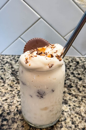 Finished Ninja SLUSHi Peanut Butter Cup Vanilla Protein Shake in a glass, topped with whipped cream, crushed peanut butter cup pieces, and a whole peanut butter cup, with a metal straw for serving.