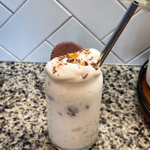 Finished Ninja SLUSHi Peanut Butter Cup Vanilla Protein Shake in a glass, topped with whipped cream, crushed peanut butter cup pieces, and a whole peanut butter cup, with a metal straw for serving.