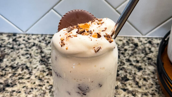 Finished Ninja SLUSHi Peanut Butter Cup Vanilla Protein Shake in a glass, topped with whipped cream, crushed peanut butter cup pieces, and a whole peanut butter cup, with a metal straw for serving.